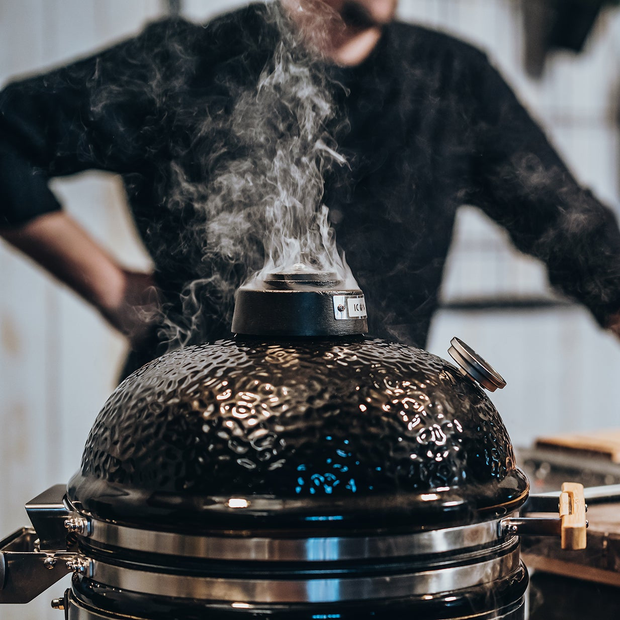 Kamado-Grill mit austretendem Rauch – Grillvorgang startet, im Hintergrund steht ein Koch bereit