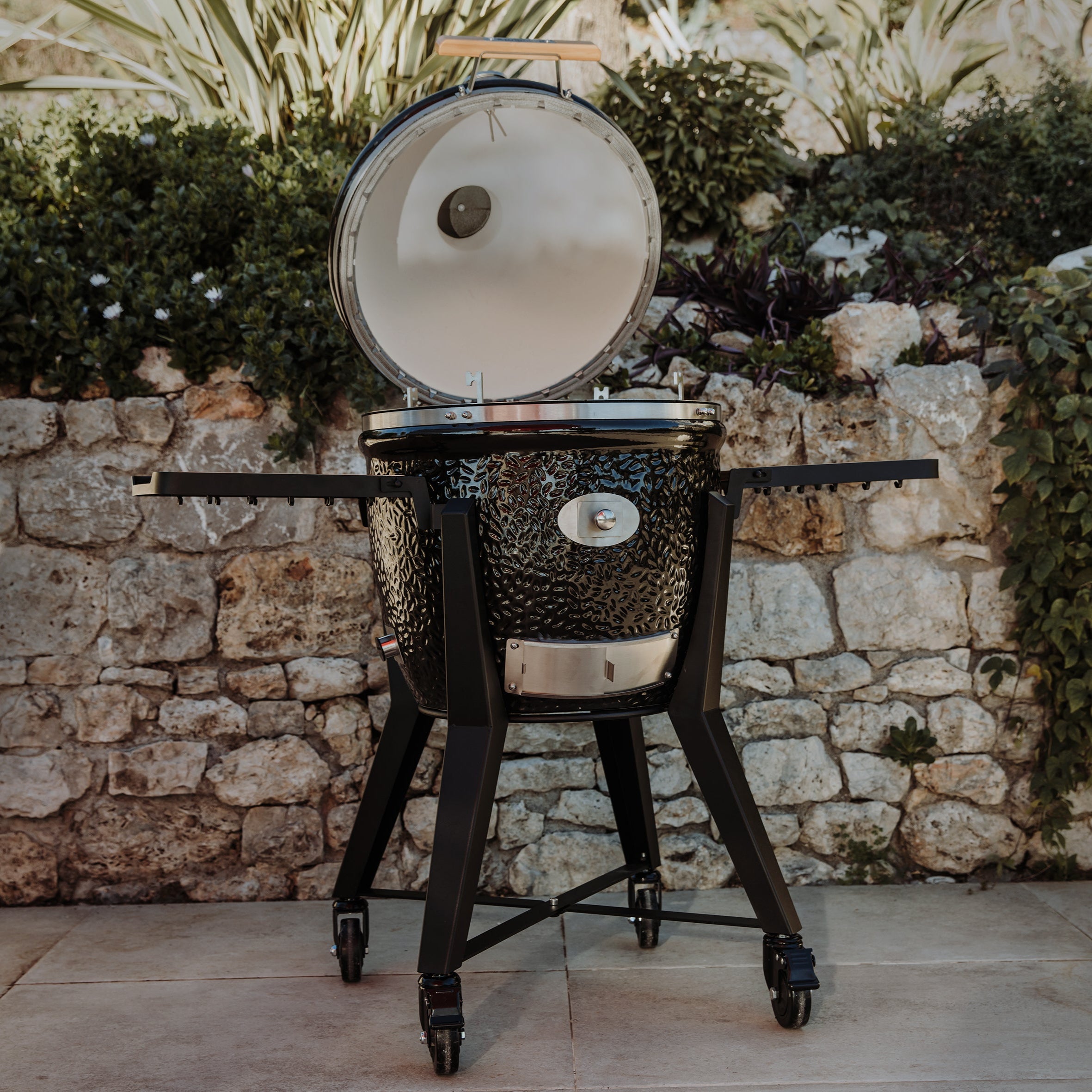 Kamado-Keramikgrill Monolith TWO 55 mit geöffnetem Deckel und ausklappbaren Ablagen, Outdoor-Szene mit Natursteinmauer und Pflanzen.
