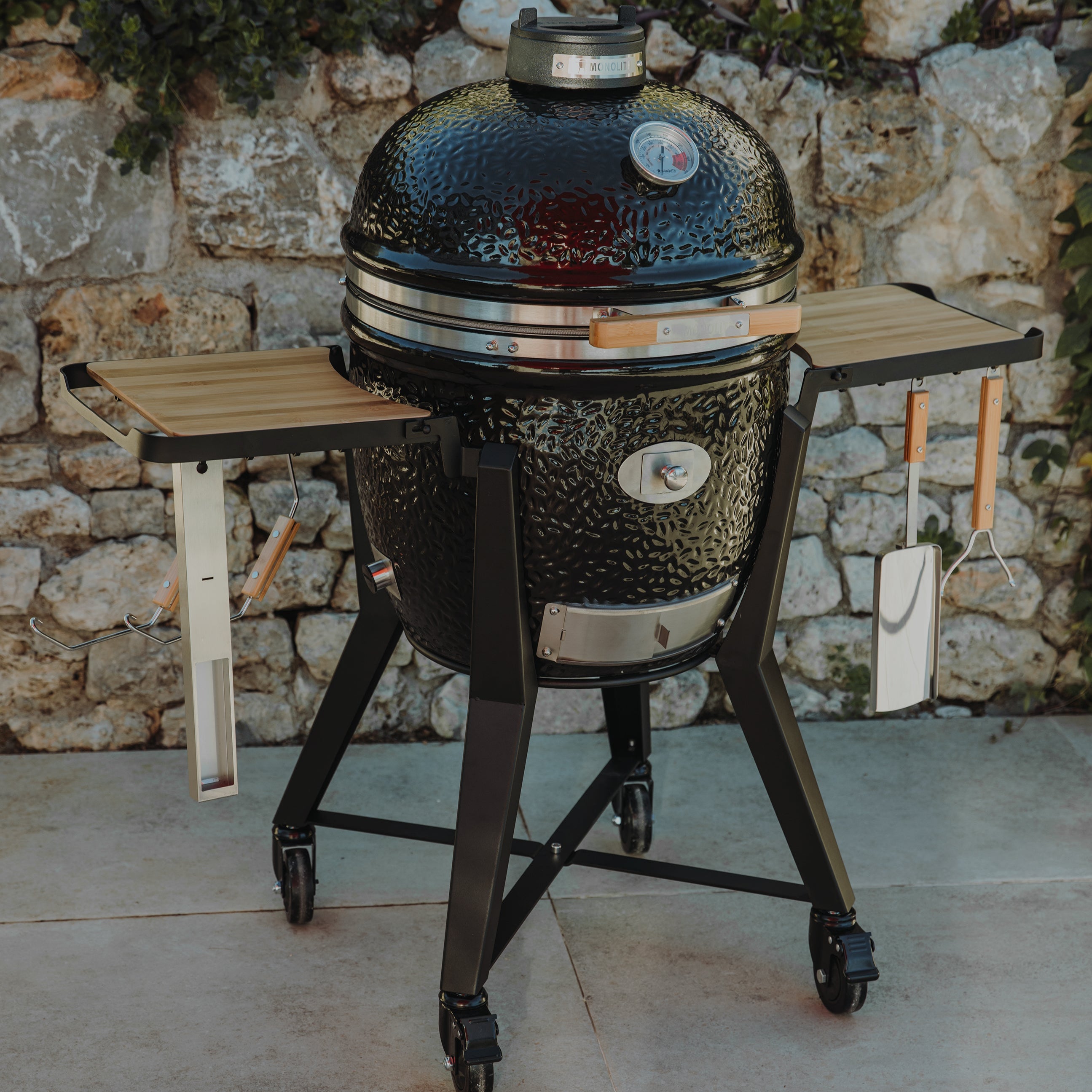 Monolith Kamado-Grill TWO 55 mit geschlossenem Deckel, Seitentischen und Zubehör, platziert auf Terrasse vor Natursteinmauer
